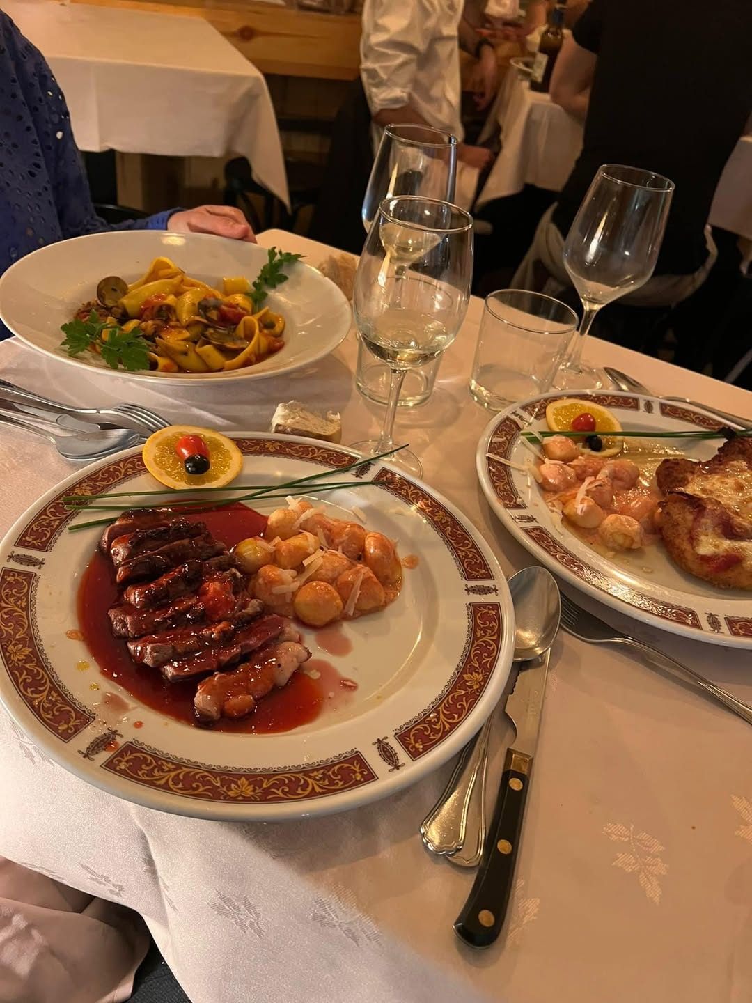 Una mesa con mantel blanco: pappardelle, pato con patatas y gambas, en vajilla clásica